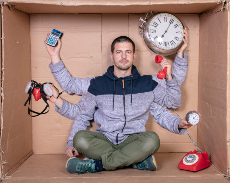 Man sitting in a box balancing a clock, a phone, a calculator and an alarm clock