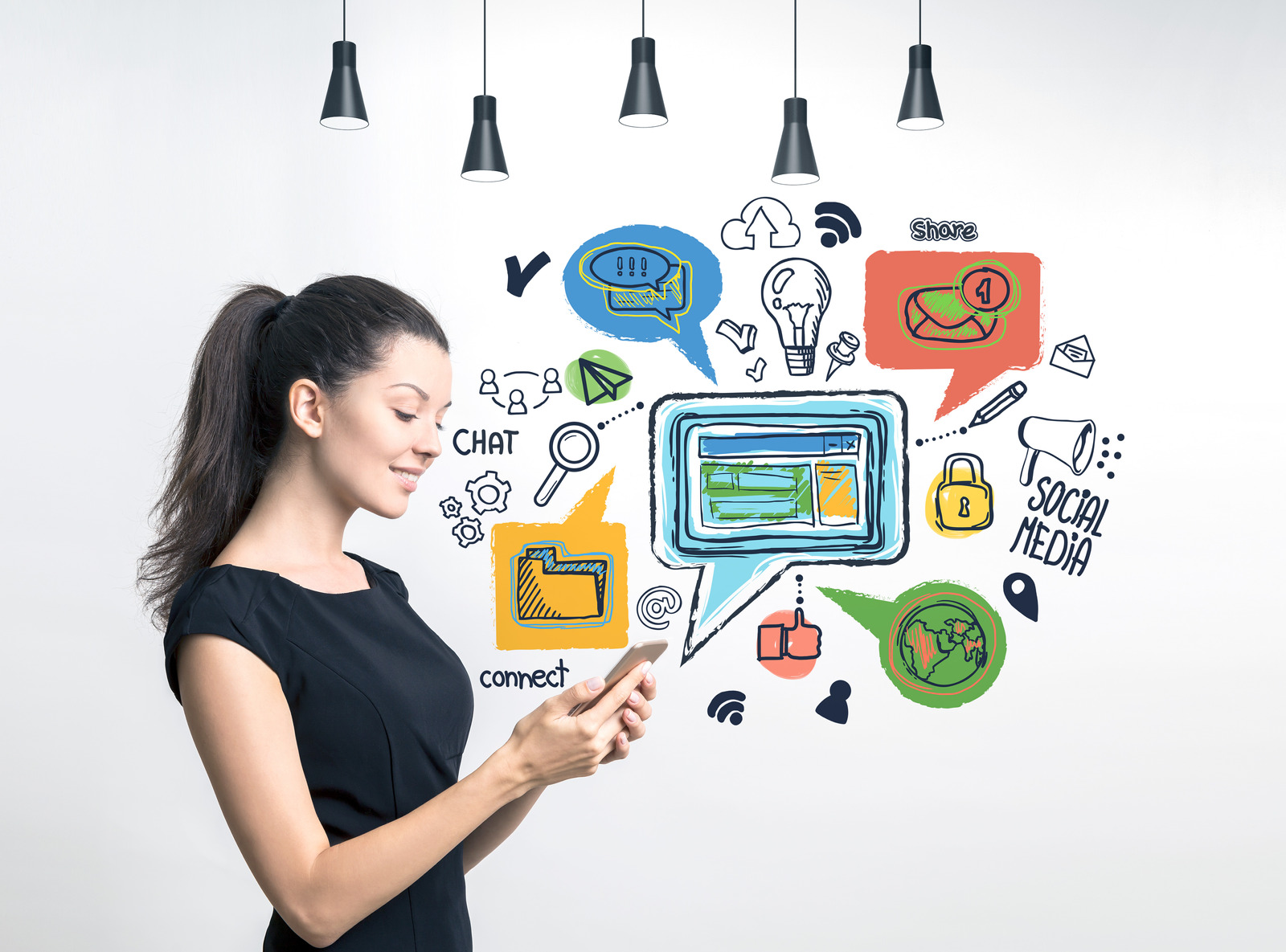 Side view of smiling young woman wearing black dress looking at her smartphone screen standing near concrete wall with colorful social media icons drawn on it.