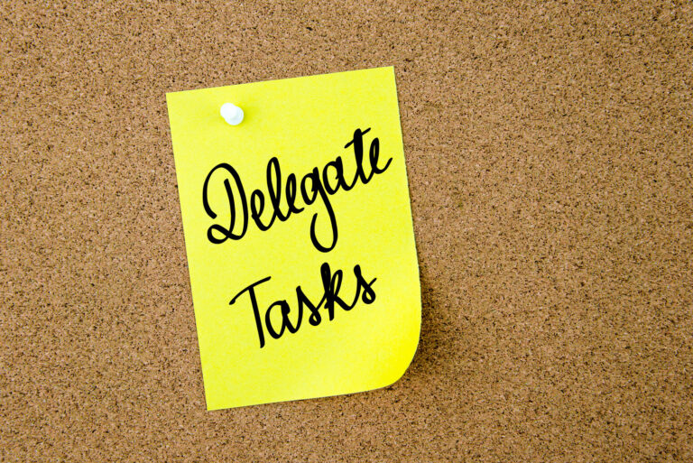 Delegate Tasks written on yellow paper note pinned on cork board with white thumbtacks, copy space available