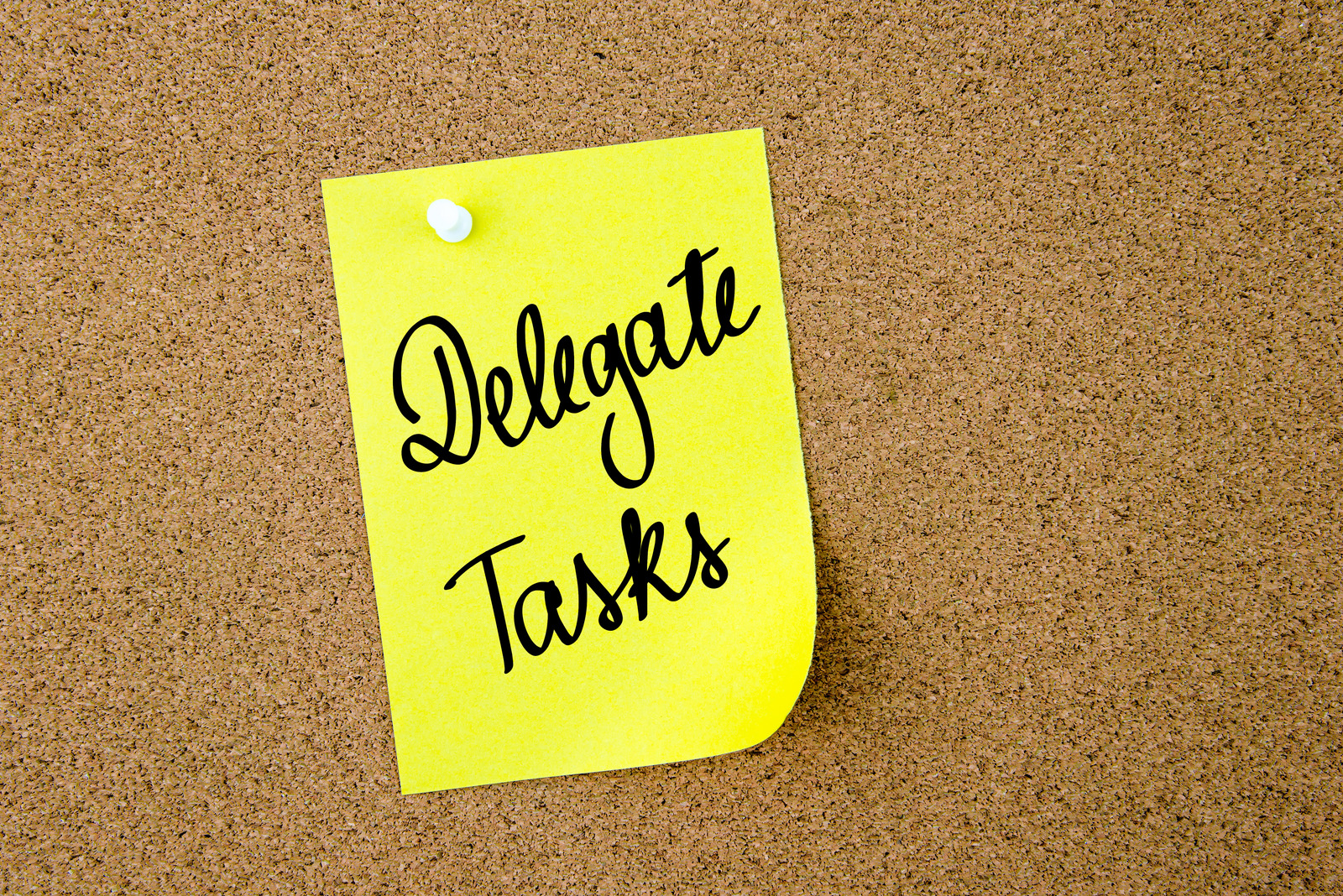 Delegate Tasks written on yellow paper note pinned on cork board with white thumbtacks, copy space available